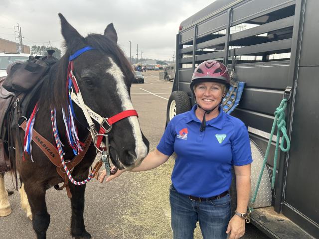 A black horse with a white streak down the center of their nose stands next to a person in a blue shirt wearing a riding helmet. 