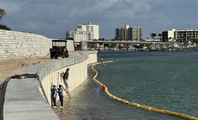 Workers in safety gear repair a waterfront barrier, with an ATV nearby and buildings lining the coast under a blue sky.