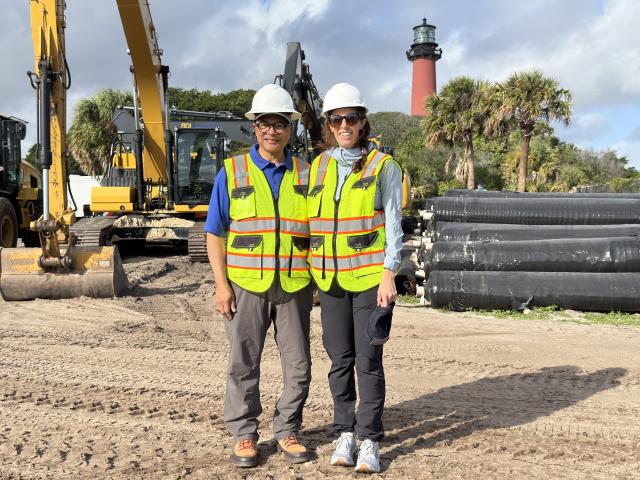 Two people in safety vests stand on a sandy site with heavy machinery and a lighthouse in the background.