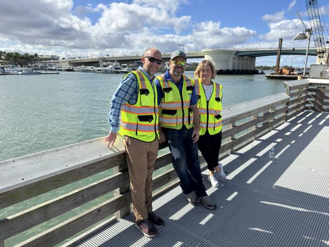Three individuals wearing safety vests stand on a dock by the water, with boats in the background and a bridge in the distance.