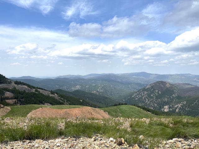 Rolling green hills and rocky terrain stretch toward distant mountain ranges under a partly cloudy blue sky. 