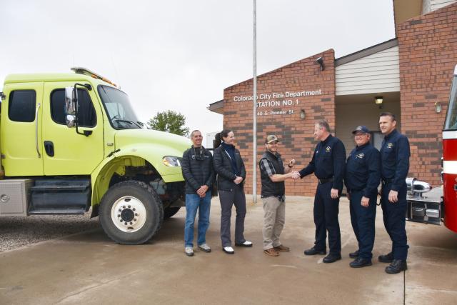 Firefighters in blue uniforms stand in front of a green fire truck while passing the keys