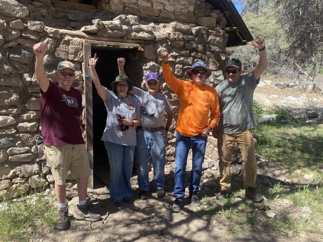 Four people stand it front of the doorway of a stone building with their arms raised in celebration.