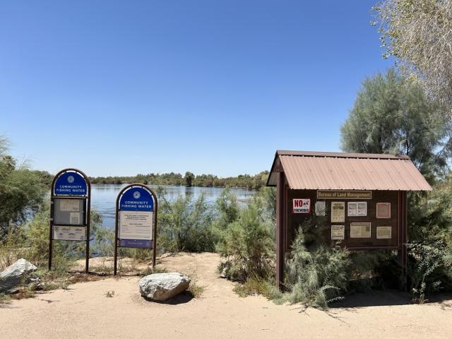 Two signs next to a calm pond with a brown wooden BLM kiosk to the right.