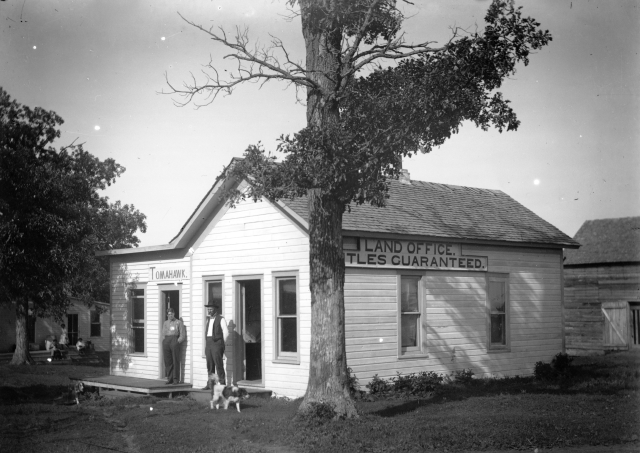 black and white photo showing two people standing in front of a small building with a dog