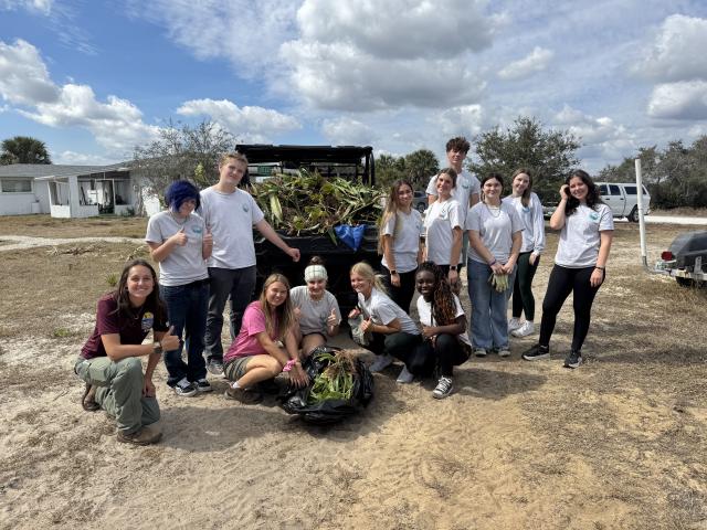 Group of young volunteers posing outdoors with collected plants and yard waste on a sunny day.