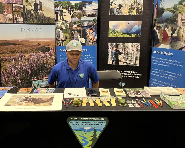 Man in blue shirt at U.S. Bureau of Land Management booth with informational pamphlets, badges, and pens on display table.