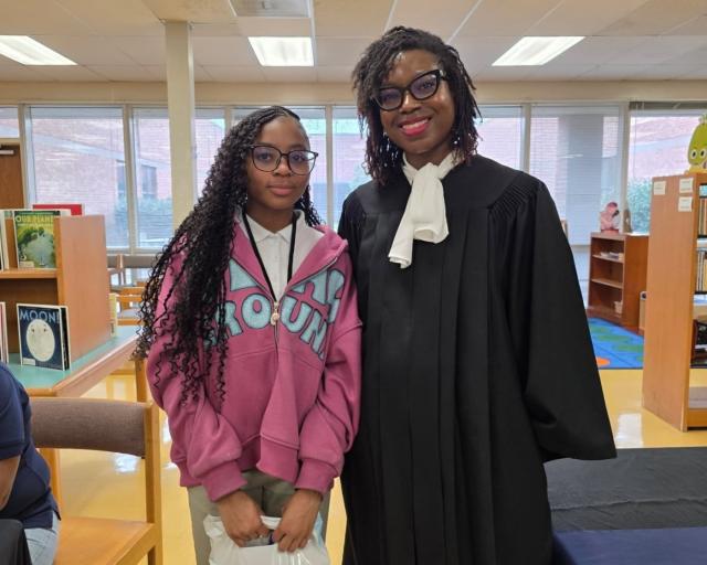 Two people standing side by side in a library, one wearing a black judge's robe and the other in a pink hoodie.