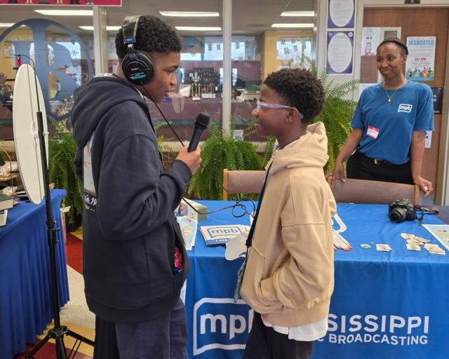 Two young people stand by a table covered with a blue "Mississippi Public Broadcasting" cloth; one holds a microphone and wears headphones.