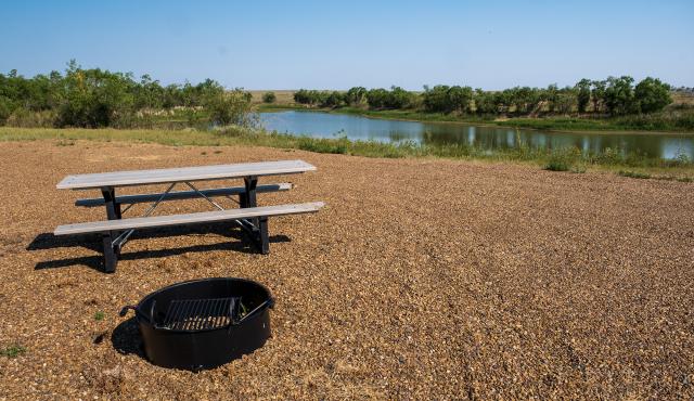 Picnic table and fire pit on gravel near a calm body of water with green trees under a clear blue sky.