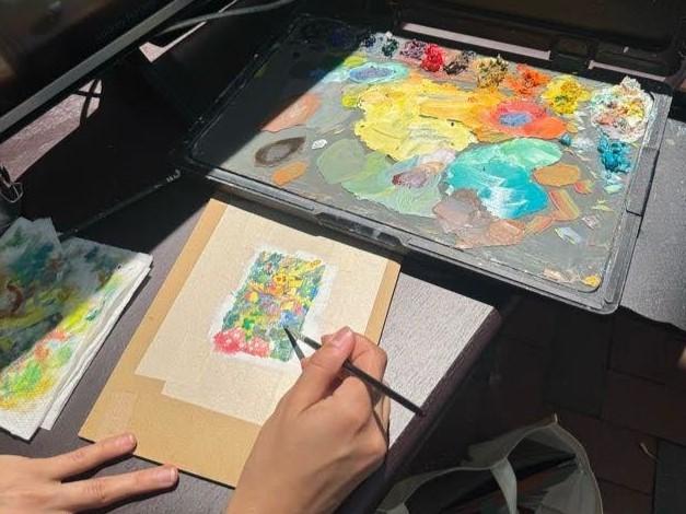 Person painting a small, colorful watercolor artwork with a brush near a palette filled with various paint colors.