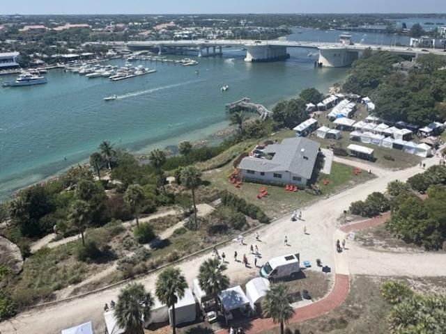 Aerial view of waterfront park with boats, bridge, tents, people walking, and greenery on a sunny day.