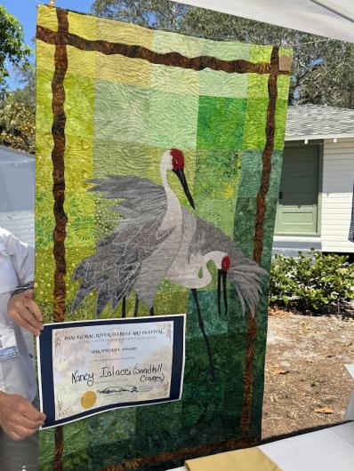 Handmade quilt depicting two sandhill cranes, held by a person also displaying a Silk Wildlife Award certificate at outdoor art festival.