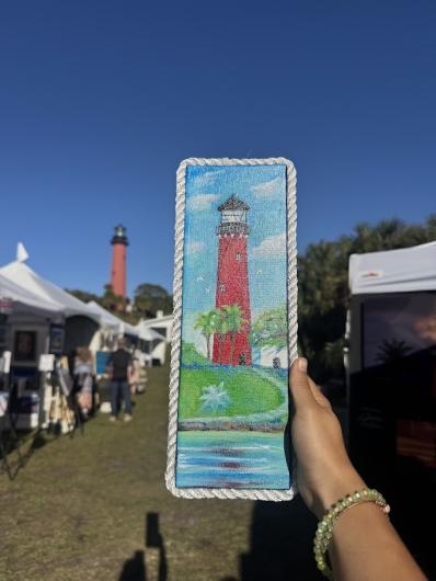 Hand holding a painted wooden panel of a red lighthouse, matching the real lighthouse in the background at an outdoor art market.