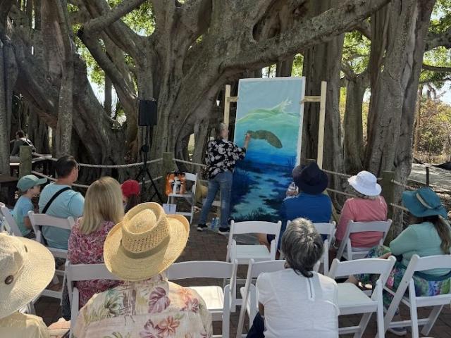 Artist painting a marine scene on a large canvas outdoors, with an audience seated on white chairs under large tree branches.