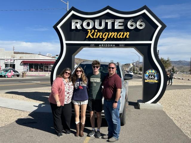 Four people stand in front of a large Route 66 Kingman roadside sign with a vintage vehicle display behind them on a sunny day.