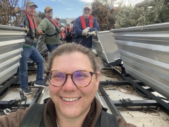 Smiling fisheries volunteer takes a selfie with other volunteers working behind her standing with loaded brush habitats.
