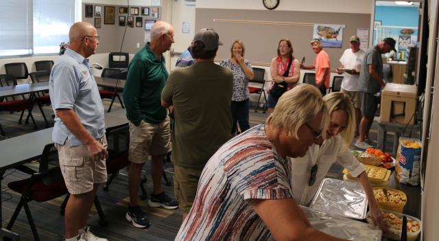 Fisheries volunteers gather in a meeting room during an appreciation luncheon, with two people preparing food at a table in the foreground.