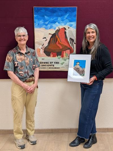 Two people stand indoors beside a colorful Canyon of the Ancients National Monument artwork, one holding a framed photo of a rocky landscape with a blue sky.