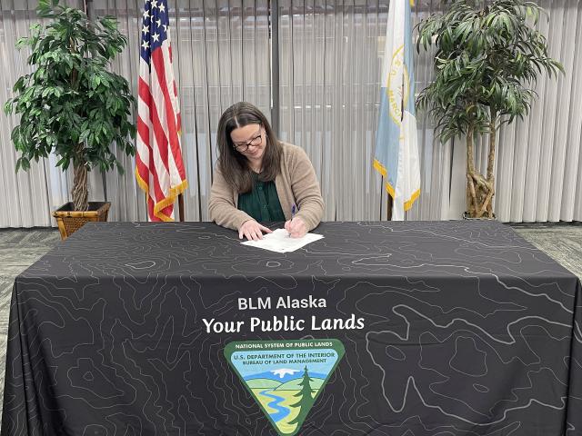 BLM Alaska Lands and Cadastral Acting Deputy State Director Chelsea Kreiner sitting at a table with a BLM table cloth and US and DOI flags behind it, signing Certificates of Allotment for three Alaska Native Vietnam-era veterans.
