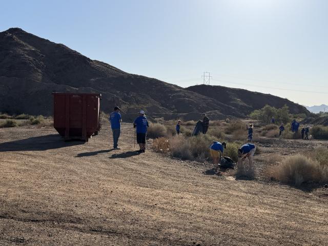 Volunteers in blue shirts collect trash from a desert wash near a large roll-off dumpster during a cleanup event on public lands near Yuma, Arizona.