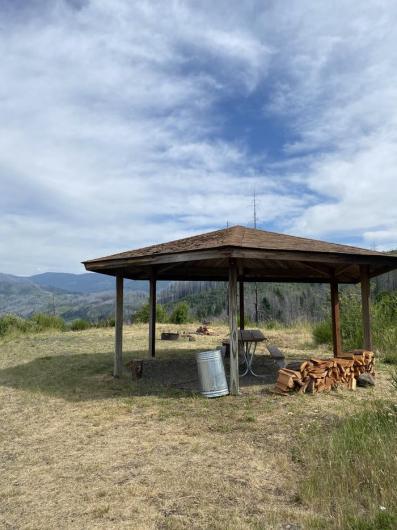 Covered wooden picnic shelter in an open grassy area with stacked firewood, a metal trash can, and distant forested mountains under a partly cloudy sky.