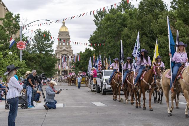 Horses and wagons and lots of people walk on a street during a parade in front of the Capitol of WY
