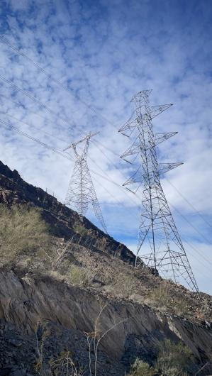 Tall transmission line towers rise along a rocky desert hillside beneath a bright, cloud‑streaked sky.