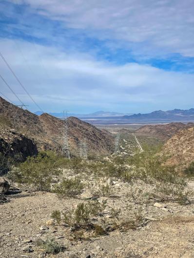 Transmission lines stretch across a rocky desert landscape with distant mountains under a partly cloudy sky.
