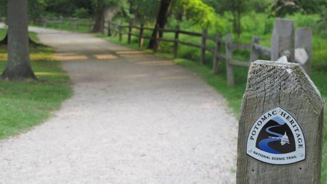 A signpost that says, "Potomac National Heritage Trail," in front of a gravel path lined with vibrant green plants