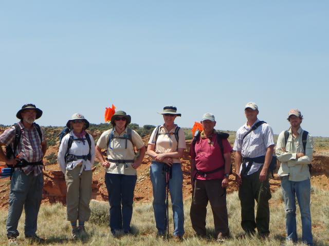 a group of people stand in a line in a grassy area with a dirt cliff in the background