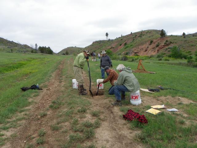 a group of four people, two with shovels, peer into a hole dug along a dirt track road