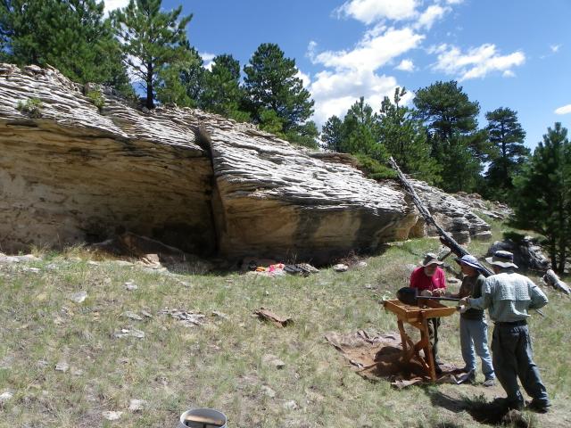 a group of three people look at a wooden stand over a hole in front of a rocky outcrop