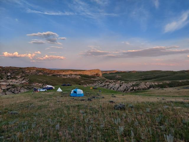 a camp with tents and vehicles sits amongst a rocky hilly area under a pink sunset