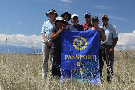 a group of people pose together in a grass field holding a flag that reads "passport in time"