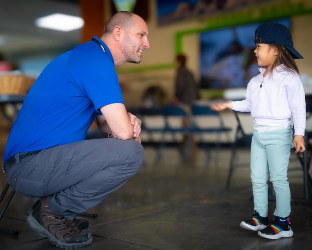 A person in a blue shirt crouches and talks with a small child wearing a backward cap indoors.