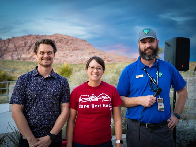 Three people stand together outdoors with desert mountains in the background; one holds a microphone.