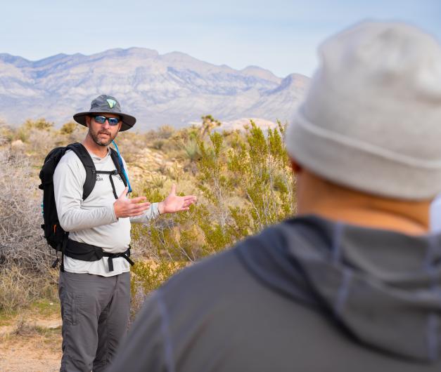 A person outdoors wearing a sun hat and backpack gestures while speaking to another person in the foreground.