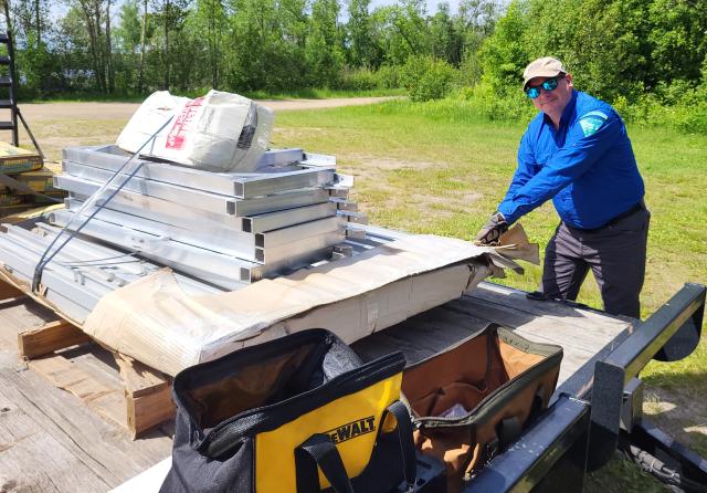 A person in outdoor work clothing stands beside a trailer, opening a large cardboard package containing metal frame components. Tool bags sit on the trailer in the foreground.