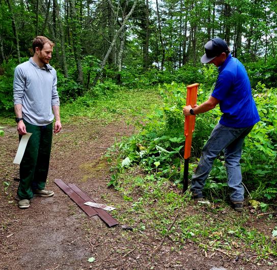 One person uses a post driver to install a trail marker stake along a forest path while another person stands nearby holding signage materials.