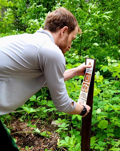 A person attaches a “Dispersed Campsite” sign to a wooden post surrounded by dense green vegetation.