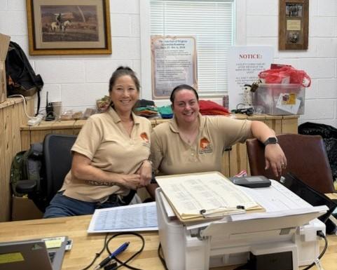 Two people sitting at a desk inside an office with rustic décor. They are smiling and wearing matching tan polo shirts with a logo. A longhorn mount hangs on the wall behind them, along with framed artwork, notices, and office supplies. A printer and paperwork are on the desk in front of them.