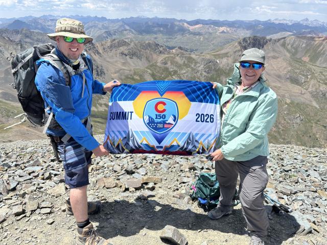 Two people stand on a rocky summit holding a flag reading "Summit 2026"