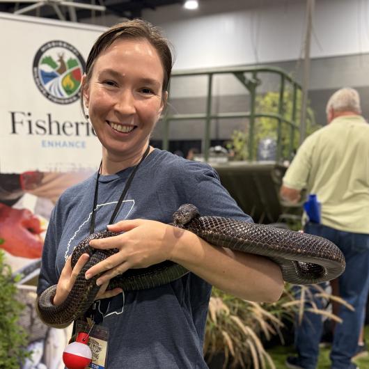 A person stands indoors holding a long dark‑colored snake across their arms. The person is smiling at the camera. A fisheries‑themed display banner and other expo equipment appear in the background, along with another attendee facing away from the camera.