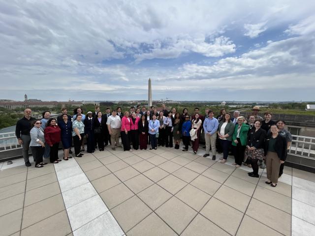 A large group of people stands together on a rooftop terrace with the Washington Monument and surrounding cityscape visible in the background under a partly cloudy sky.