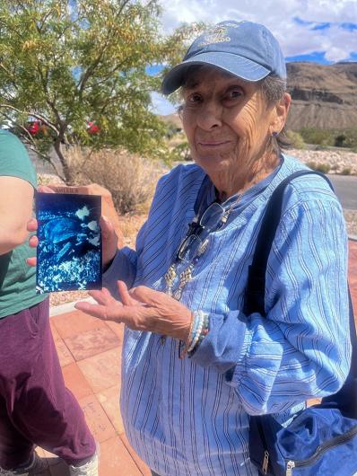 An older adult stands outdoors on a sunny day, holding up a cyanotype-style postcard with both hands. The postcard features a blue-toned image of a natural scene. The person is wearing a blue striped shirt, a blue hat, and a crossbody bag, with desert plants and rocky hills visible in the background.