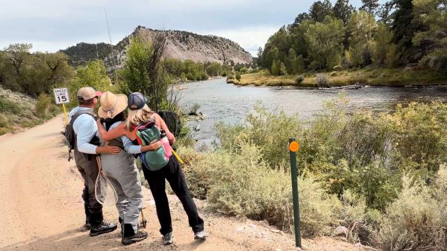 Two women and a man with their backs to the camera are looking at a peaceful river. 