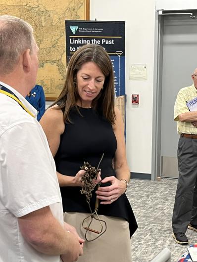 A person holds a chain surveying tool or historic mapping instrument while speaking with another person who is observing the object closely. A large wall map and interpretive display signage are visible in the background.