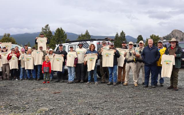 A group of people holding tan t-shirts pose for a photo outside with trees, gravel and mountains in the background under a cloudy sky.