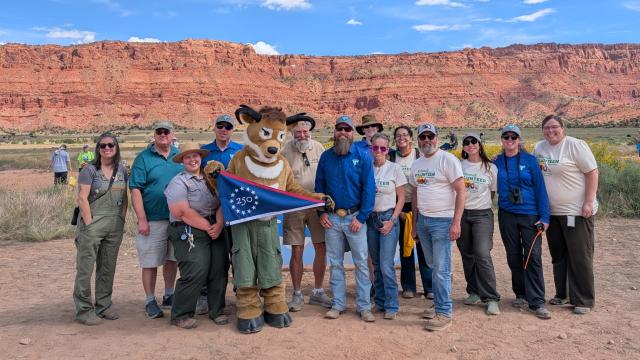 a group of people with an antelope mascot stand in front of distant red-rock cliffs